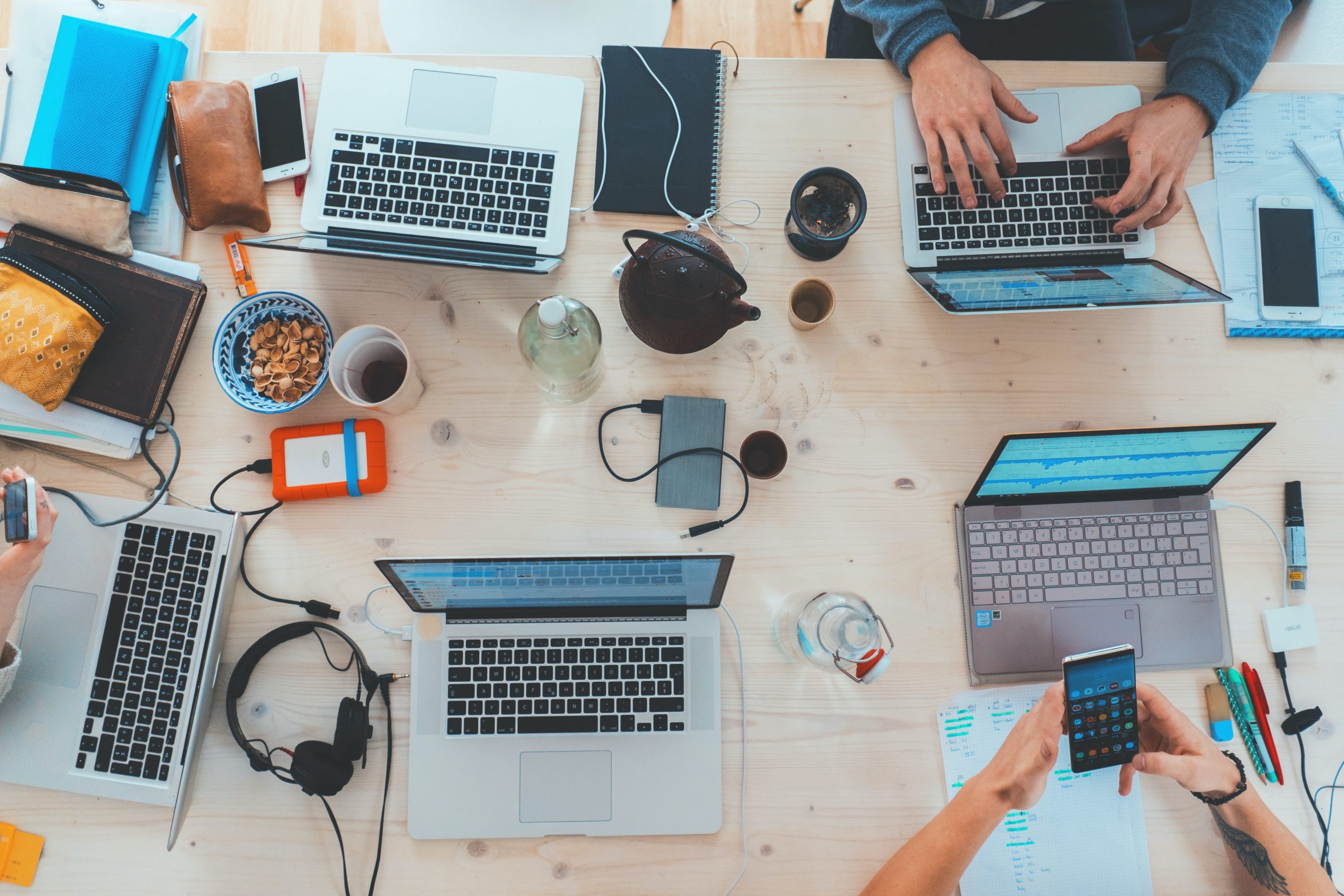 Table filled with laptops, headphone, food, pencils, and mobile devices.