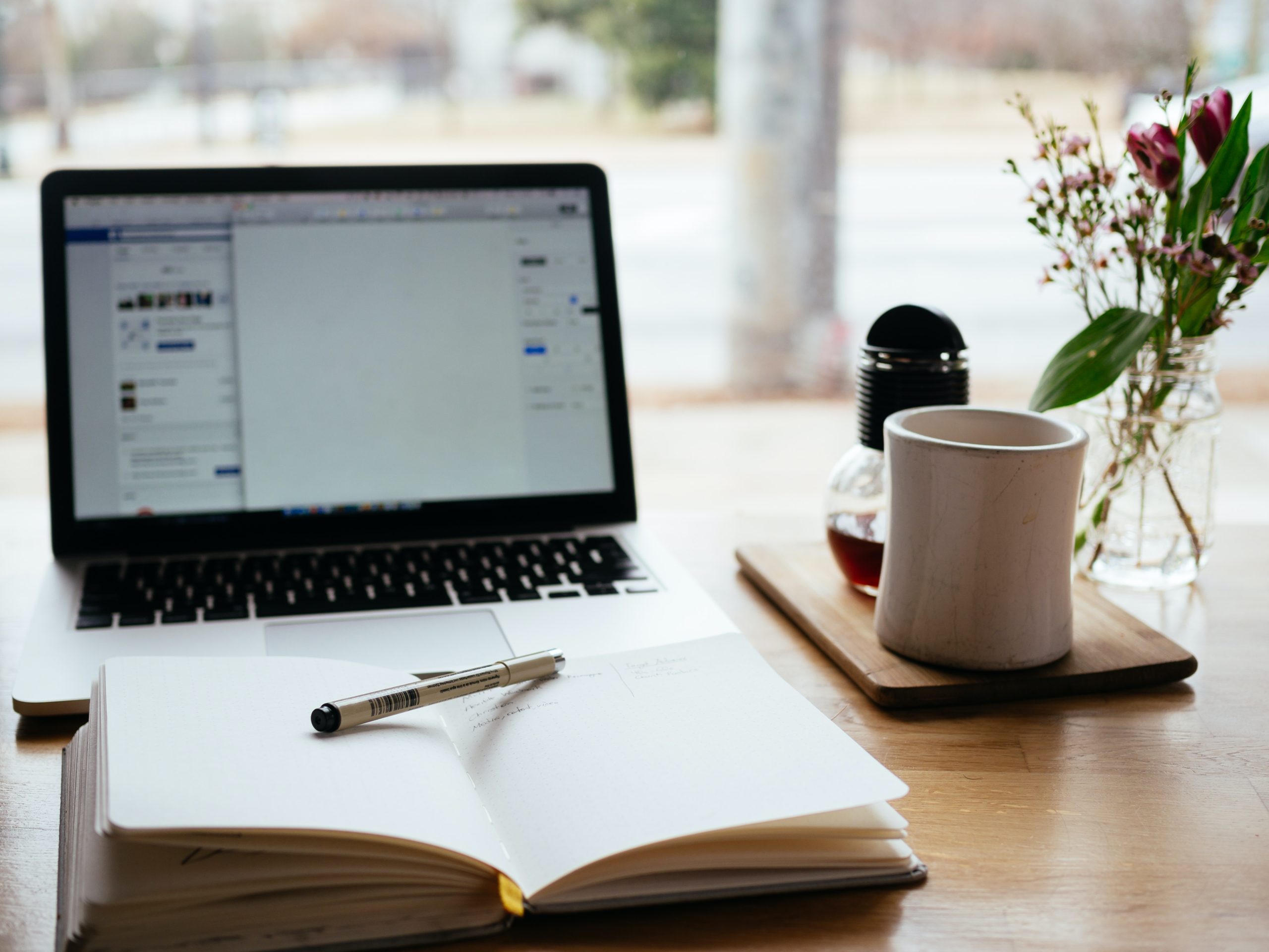 Open notebook with pen on top. Laptop blurred in background with coffee mug on desk.